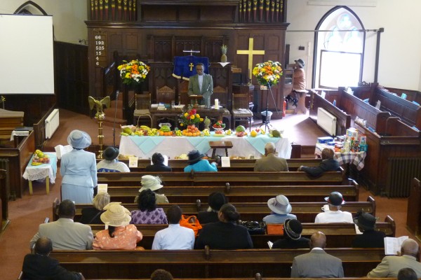 Congregation seated with one congregant standing up, a man on the stage, two tables of fruits and one table with cereal and tinned produce.